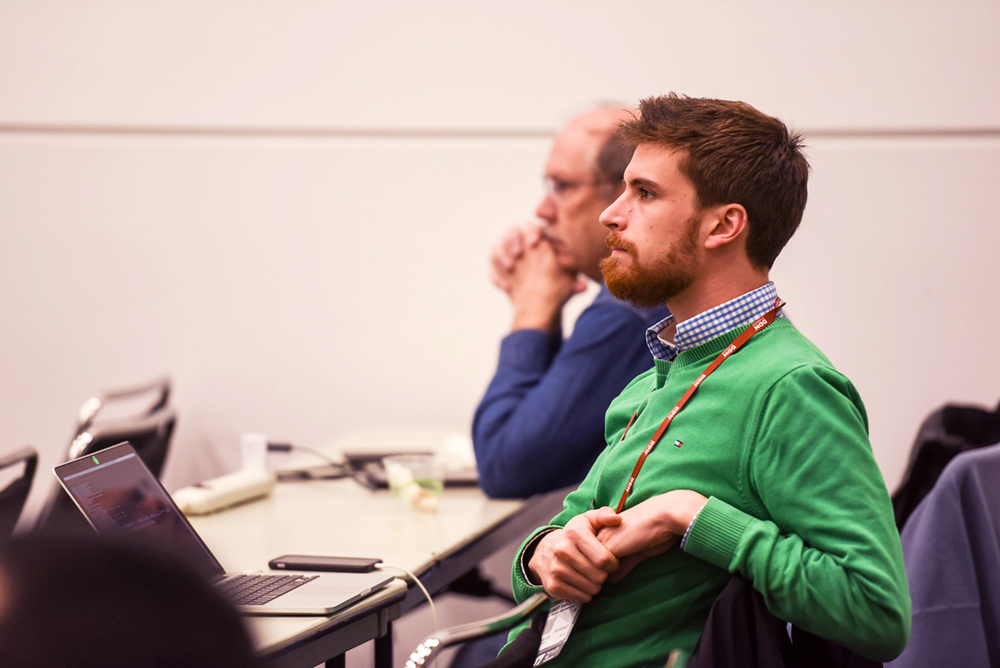 Man in a green sweater sits and listens to a presentation.